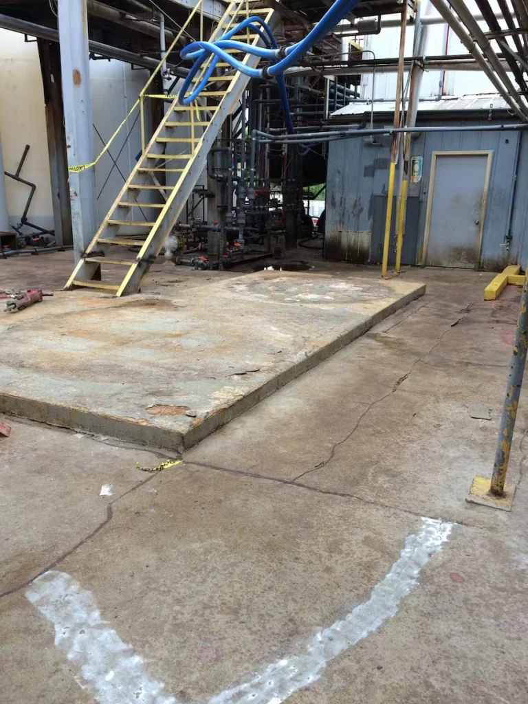 Outdoor industrial work area with a stained concrete pad and cracks in the surrounding slab; yellow metal stairs lead up to a platform, blue hoses loop overhead, and pipes and valves sit beside a small gray-sided structure with a door.