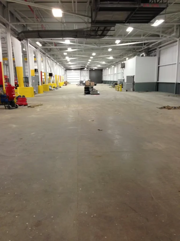 Wide view down a large industrial warehouse with a bare concrete floor, rows of white columns with yellow bases, bright overhead lights and exposed ductwork, and scattered equipment and pallets near the far end.