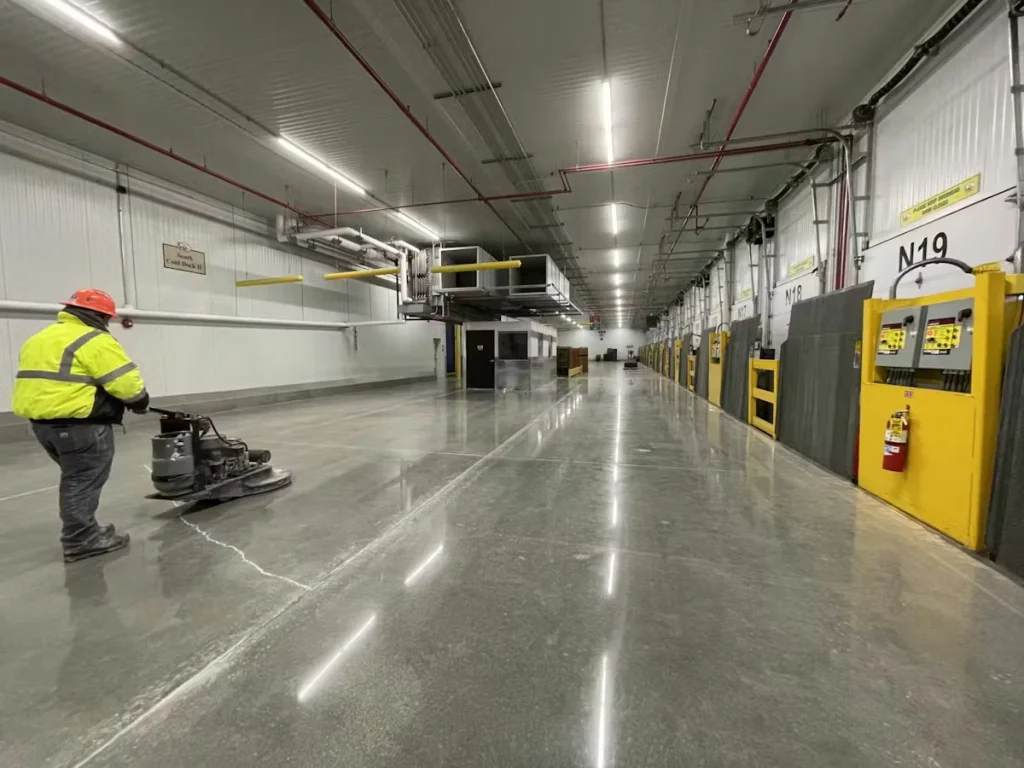 Long industrial corridor with a highly polished concrete floor reflecting ceiling lights; a worker in a high-visibility jacket and hard hat operates a floor polishing machine on the left, while numbered bay doors and yellow safety barriers line the right wall.