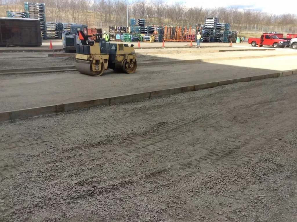 Outdoor construction yard with fresh concrete paving lanes; a small road roller compacts a gravel or base layer near the center, while orange cones, stacked materials on racks, and parked trucks sit in the background under a clear sky.