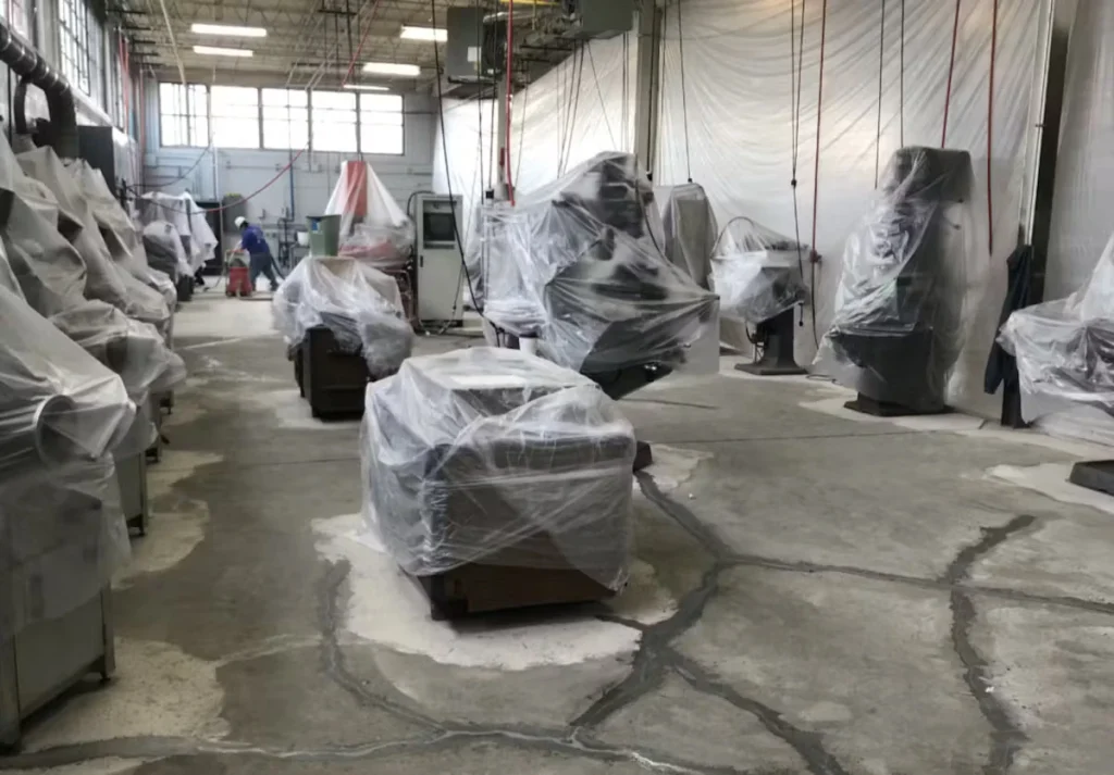 Inside a manufacturing room with many large machines covered in clear plastic sheeting; the concrete floor shows crack repairs and patch areas, with cords hanging from the ceiling and a worker in the distance near the back wall.