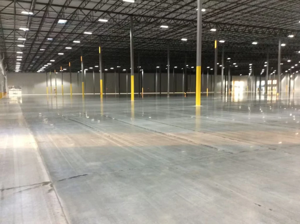 Wide view of a large empty warehouse with a glossy, freshly polished concrete floor reflecting overhead lights; rows of tall gray columns have yellow safety paint at the bases, and the dark open-truss ceiling spans the space.