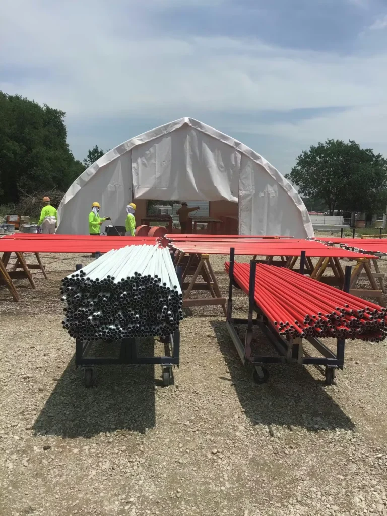 Workers in high-visibility gear stand near a white temporary tent structure; carts and racks hold bundles of red and unpainted conduit pipes in the foreground.