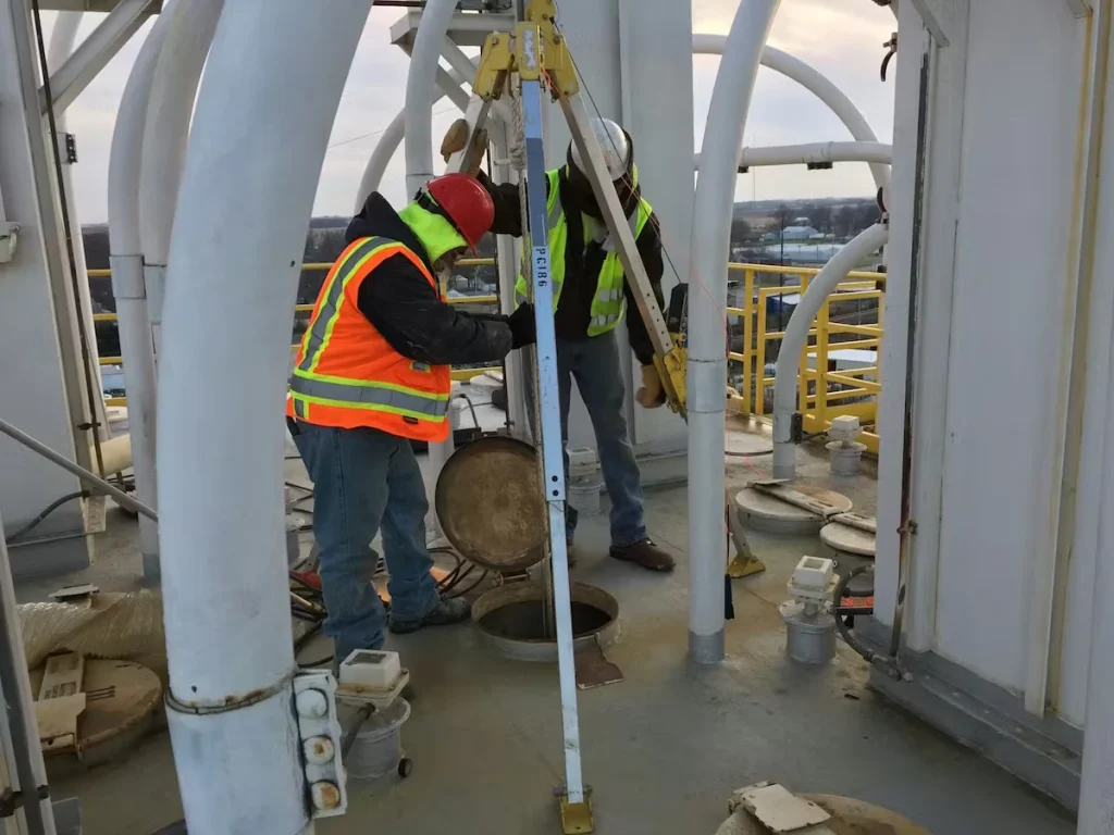 Two workers in hard hats and high-visibility vests stand on an elevated industrial platform, using a tripod hoist over an open round hatch/manhole while yellow safety railings and large white structural pipes surround them.