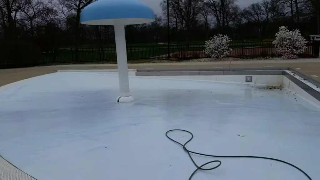 An empty outdoor swimming pool with a smooth light-colored floor; a mushroom-shaped water feature with a blue cap stands near the center, and a dark hose lies coiled across the pool bottom with trees and shrubs beyond the fence.