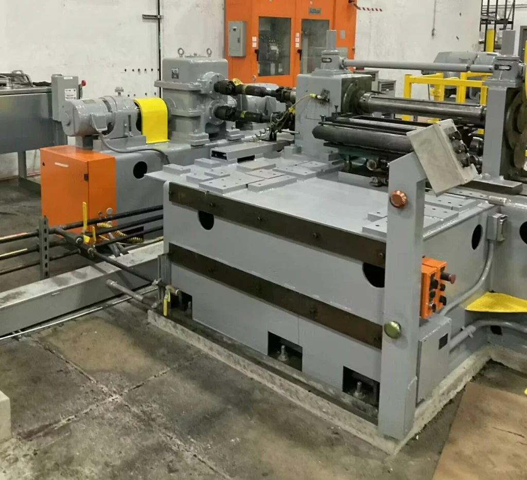 Another view of a long gray industrial machine with rollers and hydraulic components; orange panels and safety barriers nearby.