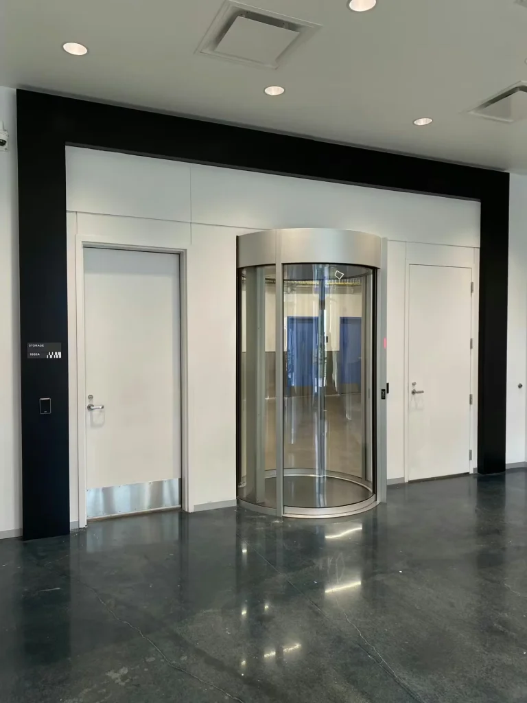Modern lobby hallway with a polished dark floor, two white doors on either side, and a central glass revolving door framed in brushed metal under recessed ceiling lights.