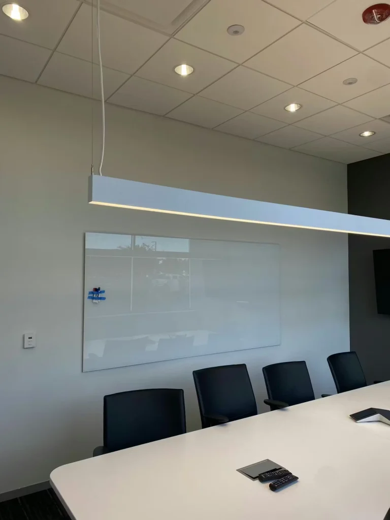 Conference room with a long white table and black chairs, a wall-mounted whiteboard, and a suspended rectangular light fixture hanging over the table.
