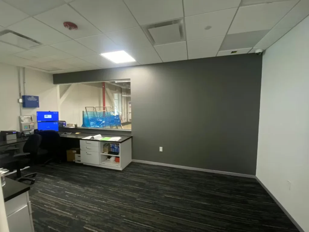 Office/control room with a large dark gray accent wall, a workstation and cabinet along the left, and a wide interior window looking into an industrial area beyond.