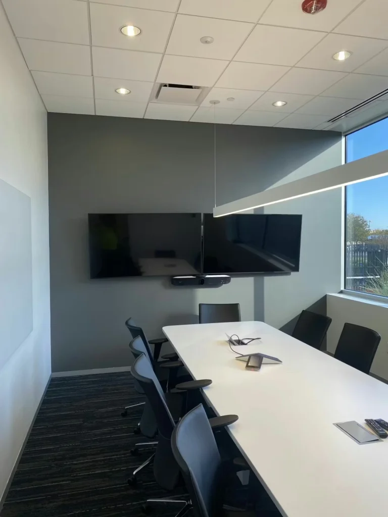 Small meeting room with a long white table and black chairs, two large wall-mounted displays above a video conferencing bar, and a corner window letting in bright daylight.