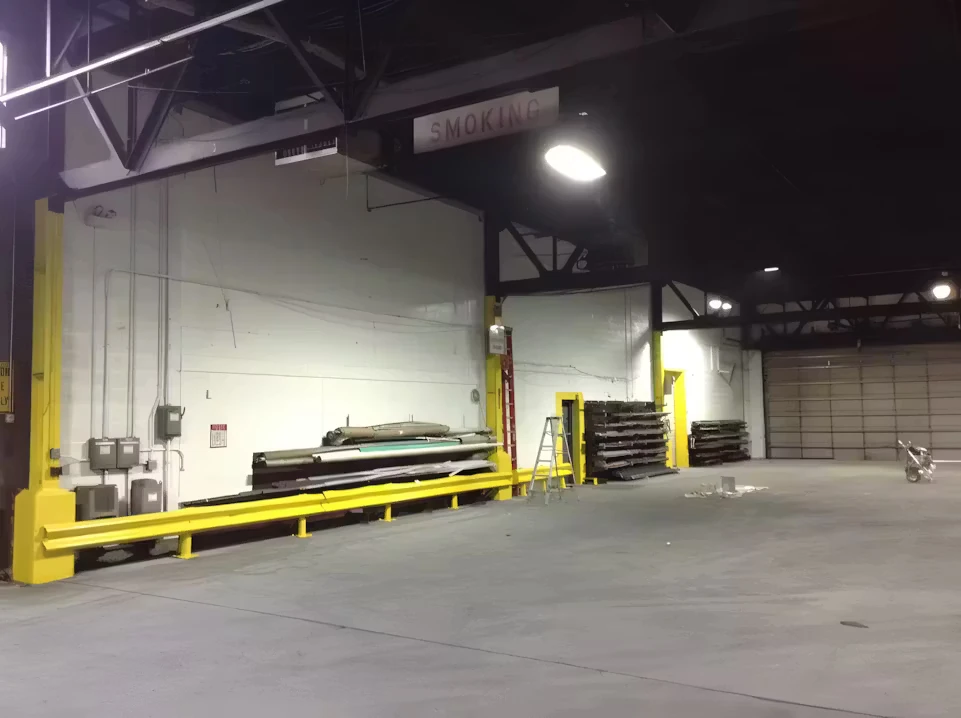 Large warehouse interior with a wide open concrete floor, white walls, yellow safety railings and column guards, stacked materials along the wall, and overhead industrial lighting with exposed beams.
