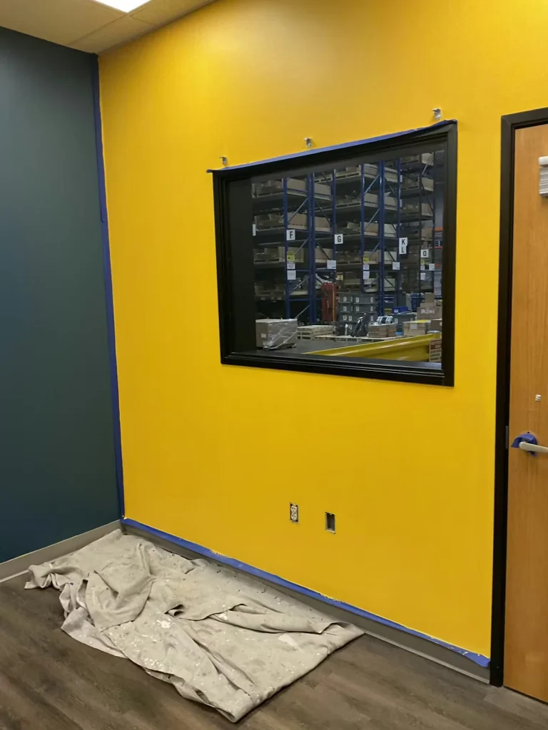 Interior room with a bright yellow painted wall bordered by blue painter’s tape, a black-framed interior window looking into a warehouse with shelving, and a wood door on the right; a drop cloth lies on the floor.