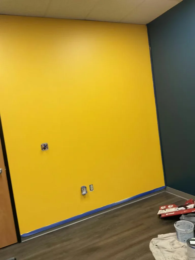 Interior corner of a room with a large freshly painted yellow wall taped along the baseboards, adjacent to a dark teal wall; exposed outlet boxes and painting supplies sit on the floor.