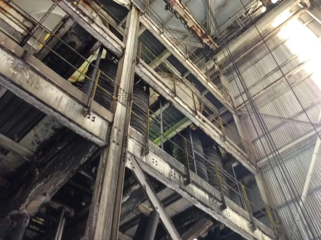 Upward view inside an industrial facility showing a tall steel structure with catwalks and railings, heavy grime and rust on beams, large cylindrical equipment, and a high corrugated metal roof with hanging cables.