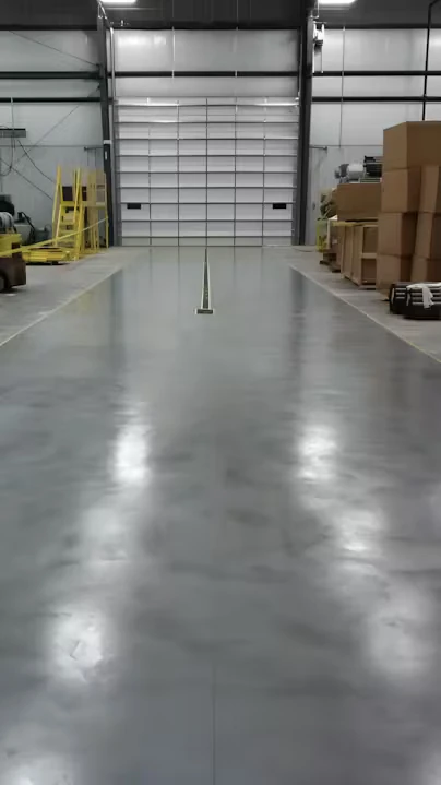 Warehouse interior with a shiny gray epoxy floor leading to a large roll-up door; a narrow recessed strip runs down the center, with pallets and stacked boxes along the right and equipment and safety barriers on the left.