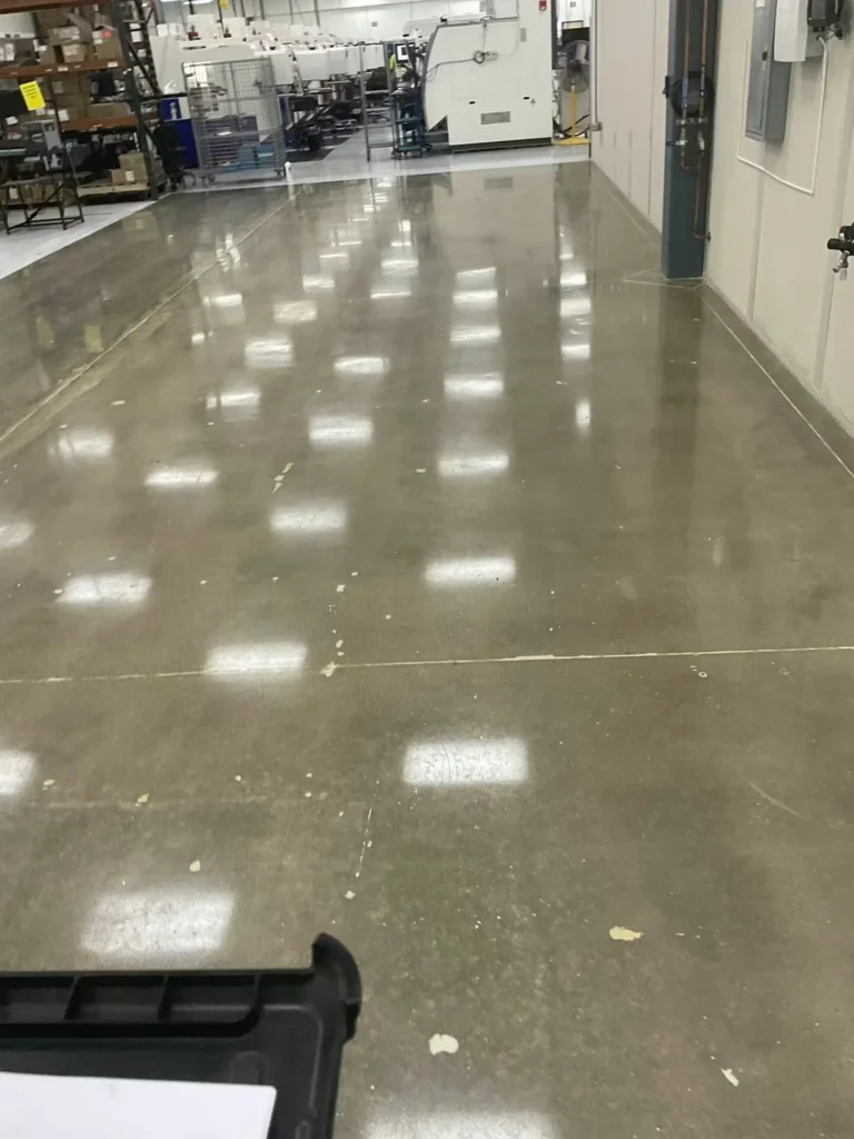 Polished concrete floor in an industrial workspace, reflecting overhead lights; machinery and storage racks line the sides near the back of the room.
