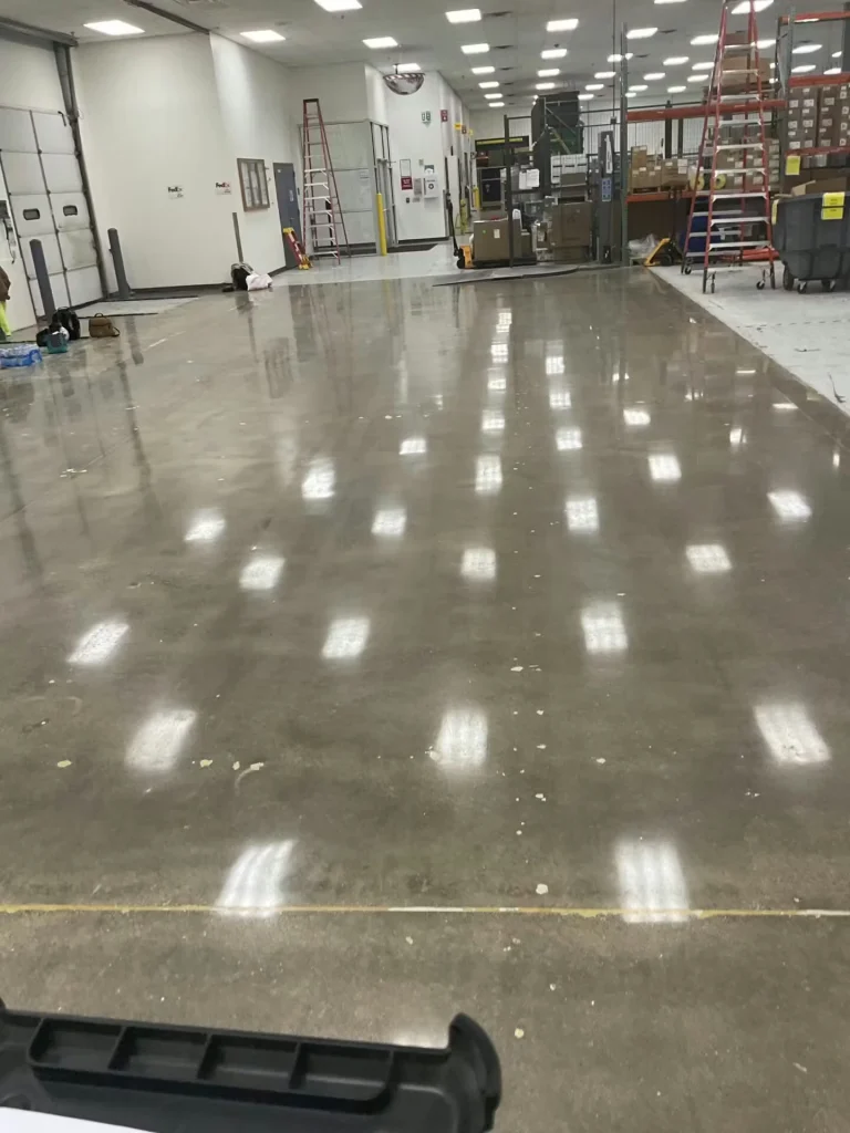 Wide view of a glossy concrete floor inside a warehouse area, with shelving, boxes, and a large green baler or compactor near a roll-up door.