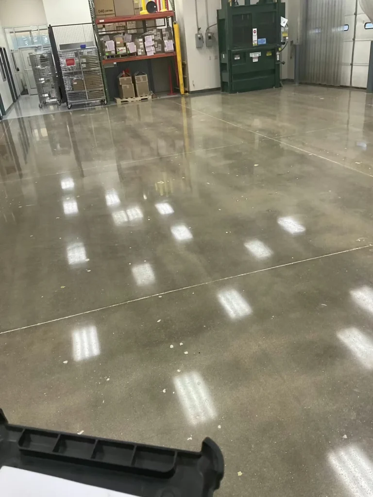 Large warehouse interior with a freshly polished concrete floor reflecting ceiling lights; ladders, pallets, and stacked boxes sit along the perimeter