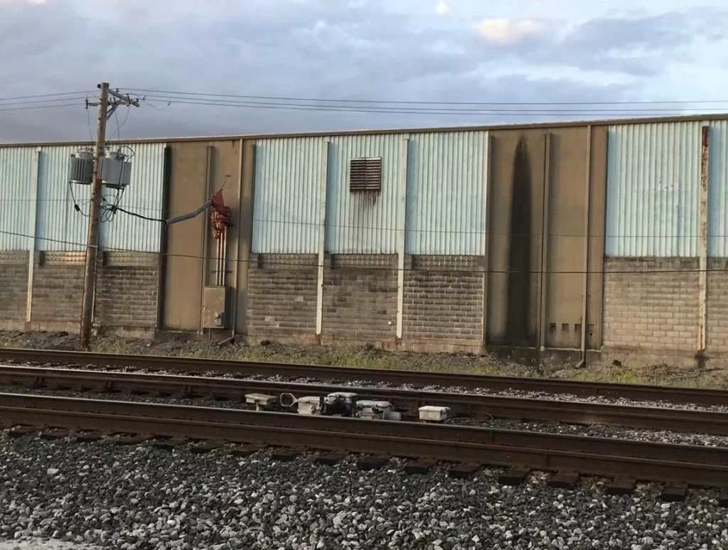 Exterior of an industrial building with corrugated metal siding and a cinderblock base, viewed across railroad tracks; a utility pole and power lines stand at left, and a dark vertical stain marks one section of the wall.