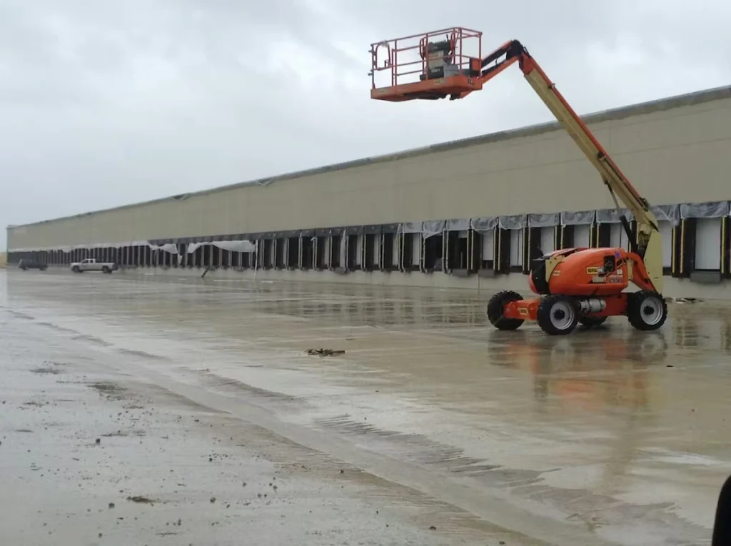 Rainy exterior of a long distribution center with many loading dock doors; an orange boom lift is extended near the building over a wet concrete lot.
