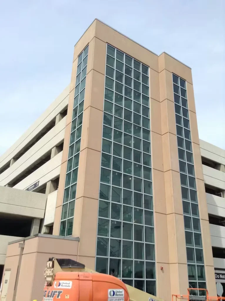Tall glass stairwell tower attached to a multi-level concrete parking garage, viewed from below; an orange boom lift sits at the base near the building.
