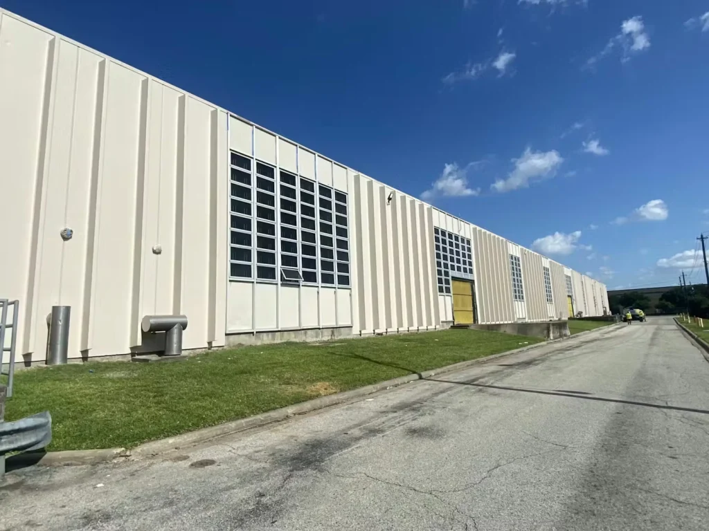 Long light-colored industrial building under a bright blue sky, with a grass strip and roadway running alongside.