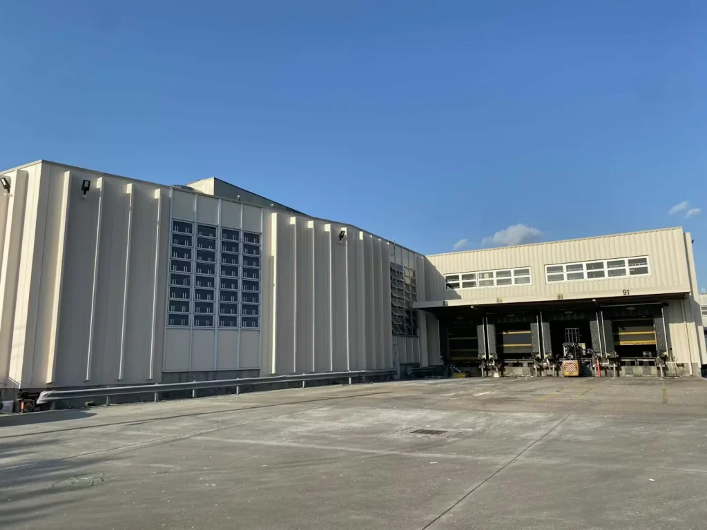 Angled view along a warehouse exterior wall with vertical metal panels, strong sunlight, and long perspective down the roadway.