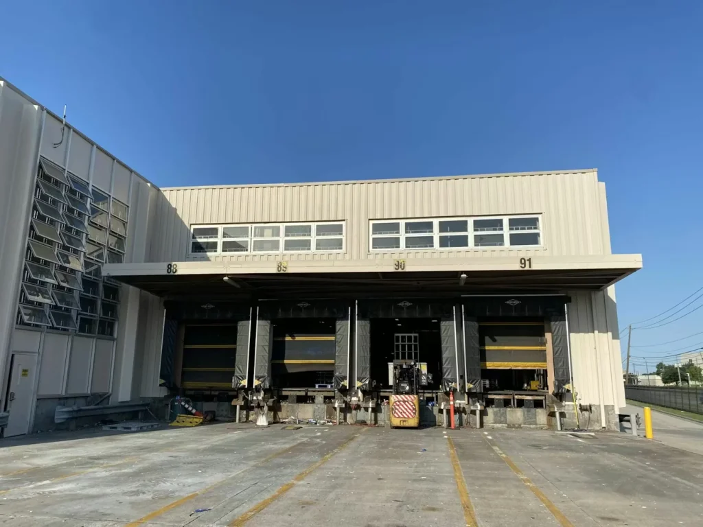 Long warehouse wall with louvered window sections and a narrow grass verge, shot at an angle under a clear sky.