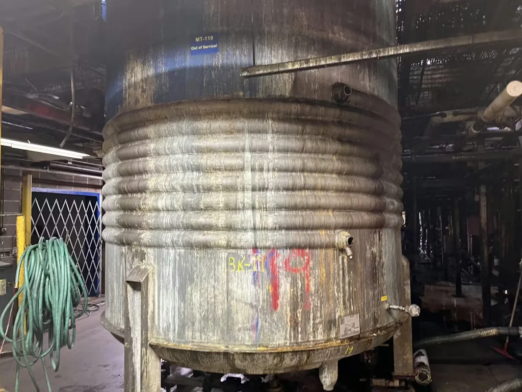 Large grimy industrial tank with coiled piping and hoses in a dim factory area.