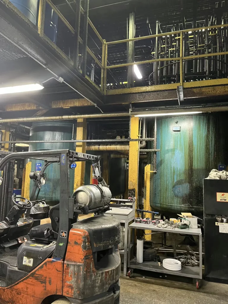 Industrial plant interior with yellow catwalks, large process tanks, and drums on the floor below.