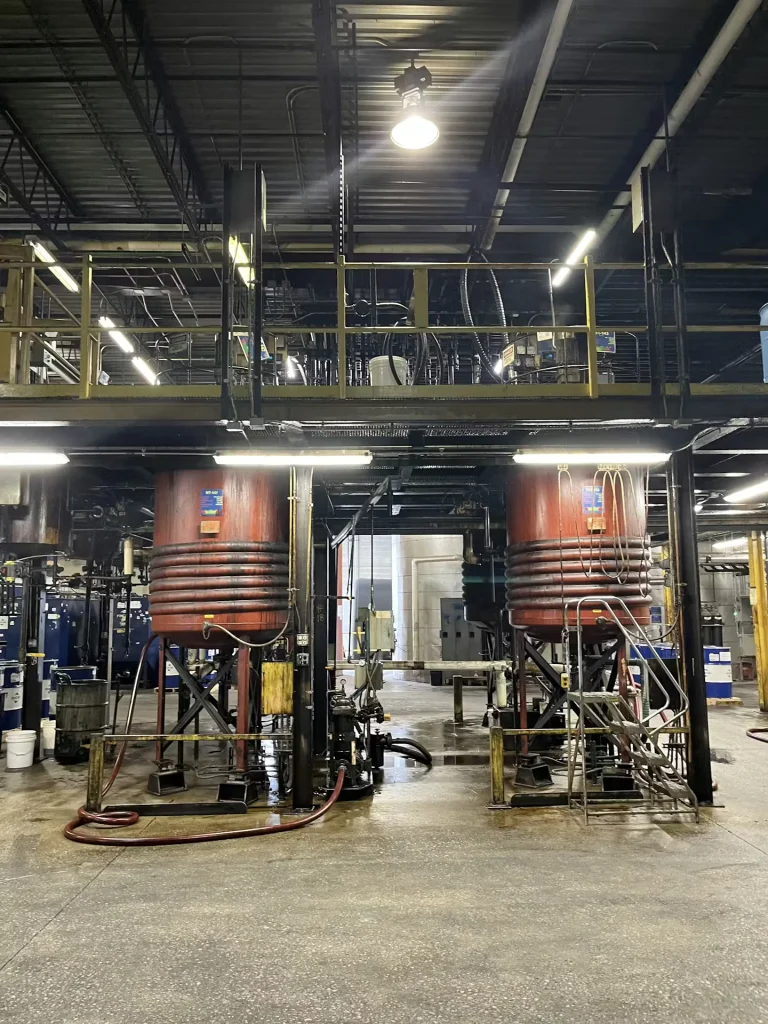 Wide view of an industrial floor with two large red tanks under a mezzanine and hoses across a wet concrete surface.