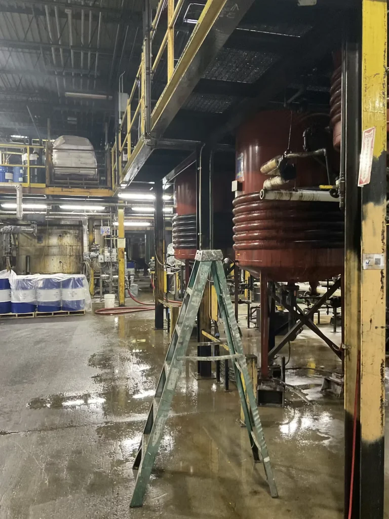 Industrial work area with a ladder in the foreground, puddled floor, and large red process tanks beneath overhead catwalks.