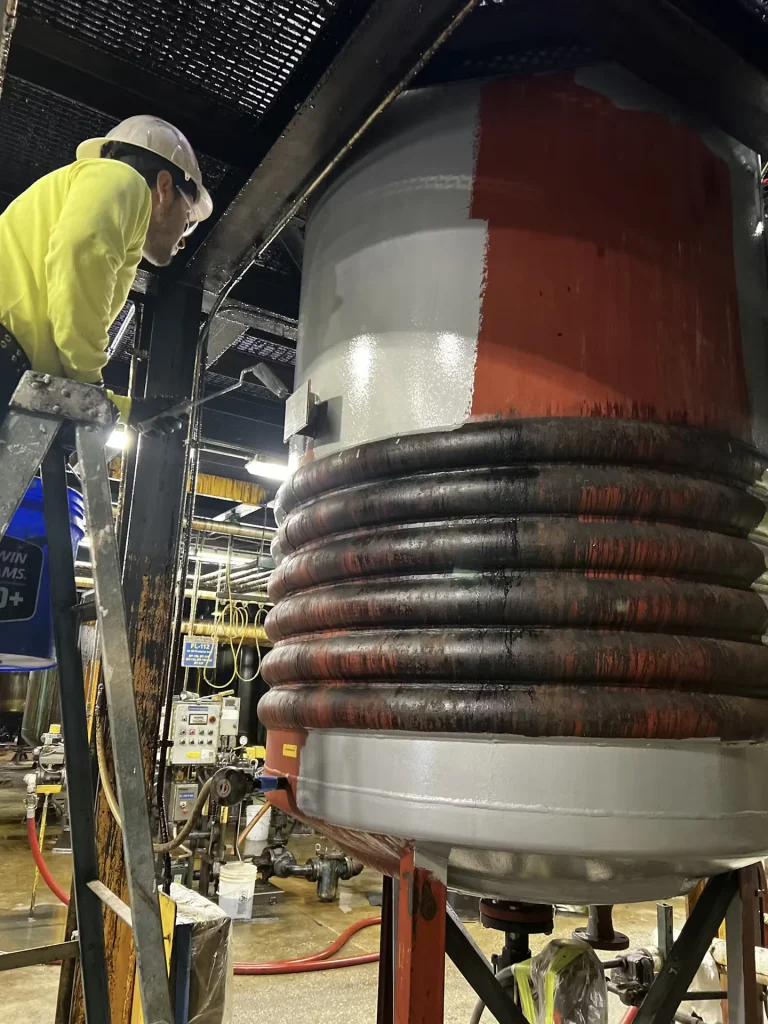 Worker in a hard hat using a roller to repaint a large tank, with fresh gray coating over older red paint.