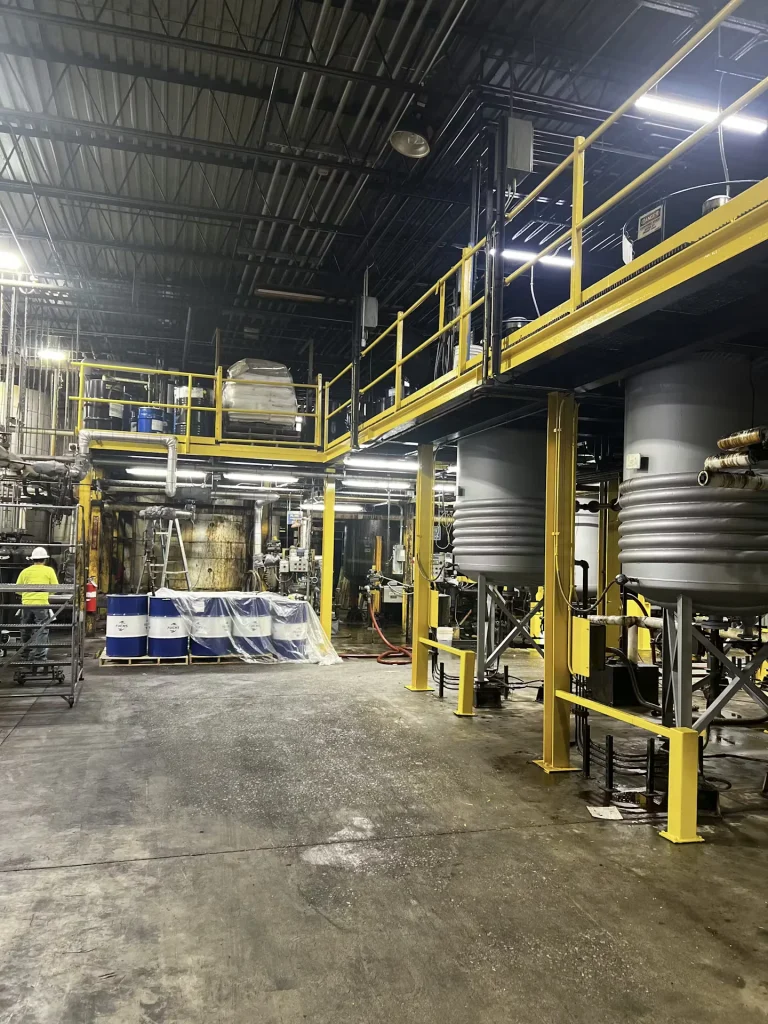 Industrial facility with yellow-railed catwalks above large process tanks and equipment arranged on the factory floor.