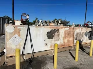 Rust-stained rectangular fuel tank outdoors, surrounded by yellow bollards and hoses, before repainting.