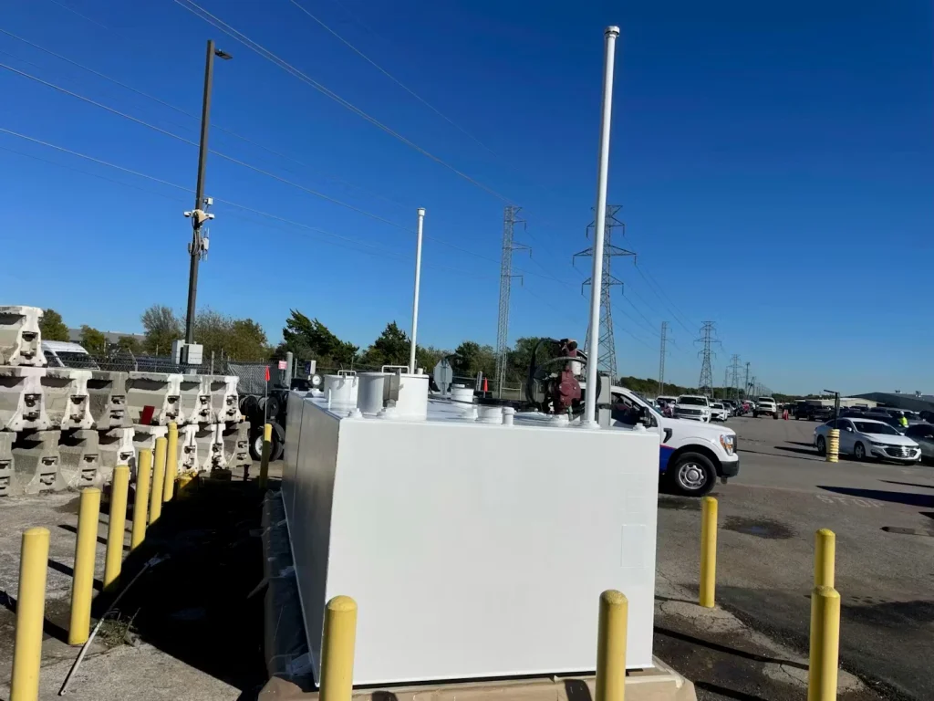 End view of a freshly painted white fuel tank with tall vent pipes, set in a lot near power lines and parked vehicles.