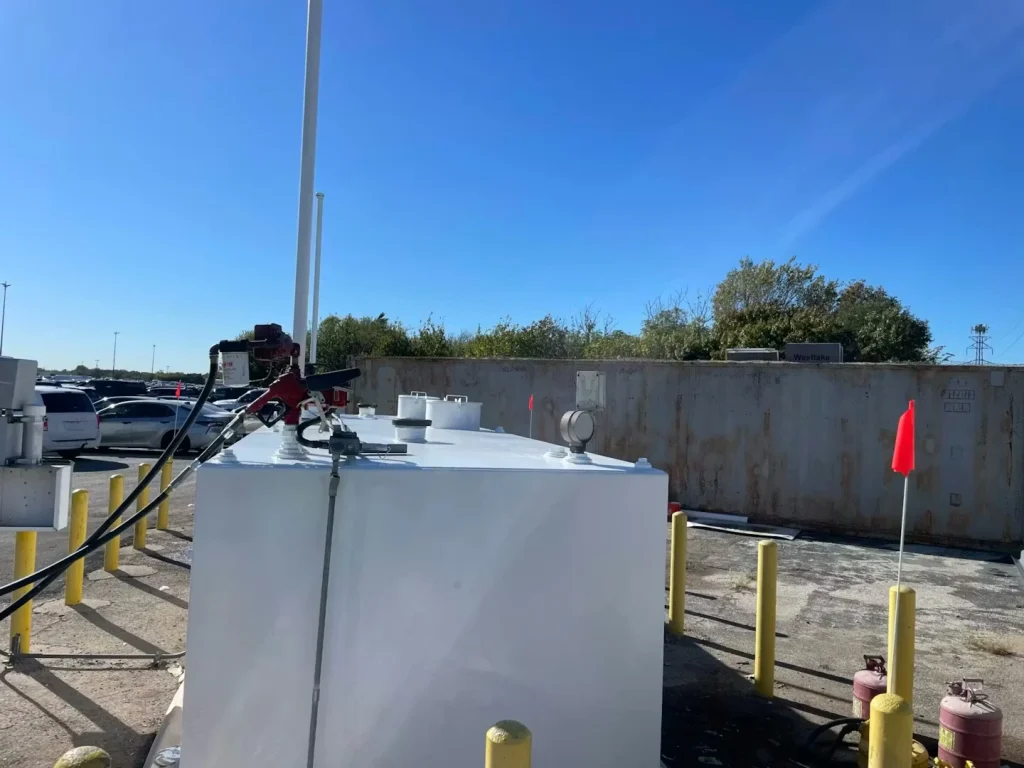 Freshly painted white rectangular fuel tank outdoors with a fuel pump and hoses on top, surrounded by yellow safety bollards under a clear blue sky.