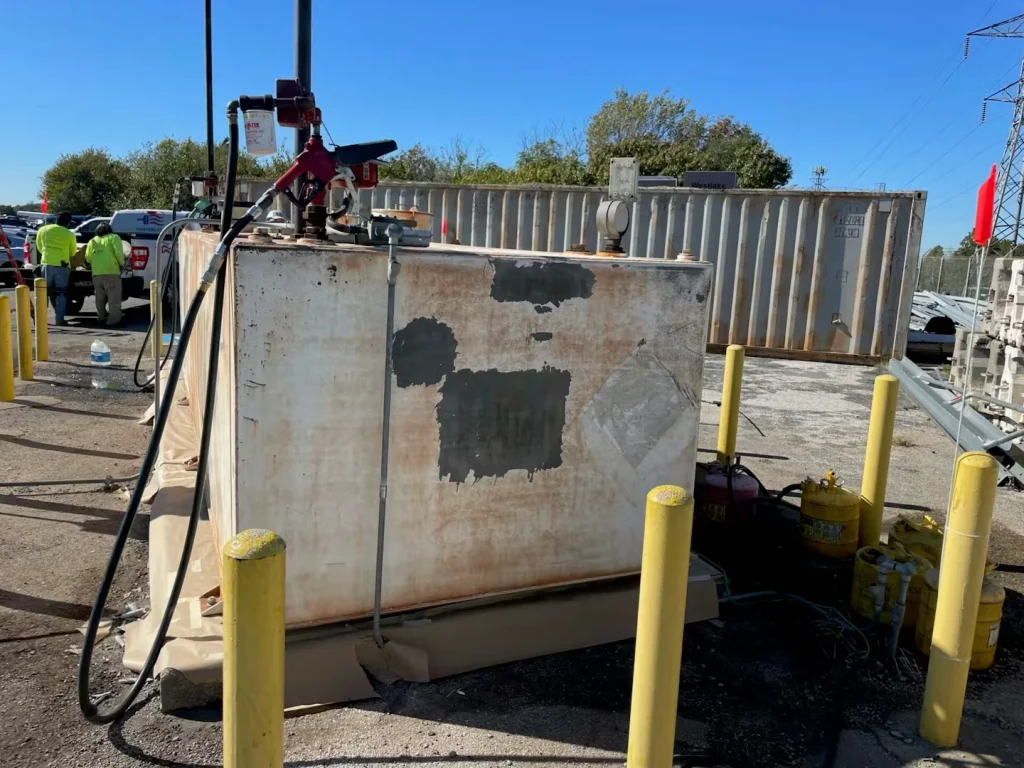 Weathered rectangular fuel tank with patchy primer and rust stains, fuel hoses and equipment on top, set among yellow bollards in a paved lot.