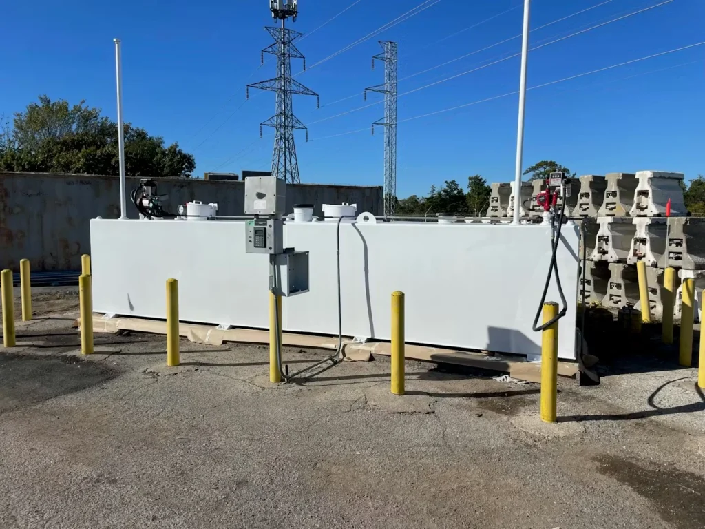 Long white-coated fuel storage tank in an outdoor lot with a mounted control box and hanging fuel hose, lined with yellow bollards and concrete barriers beneath power lines.