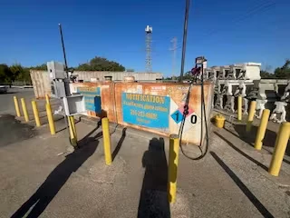 Front view of a weathered fuel tank with a “NOTIFICATION” sign, hoses, and yellow bollards in a paved yard.