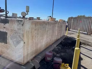 Side view of a corroded fuel tank with paint cans and supplies underneath, enclosed by yellow bollards.