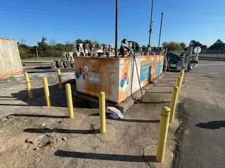 Wide shot of a rusted fuel tank installation in a parking area, ringed by yellow safety bollards.