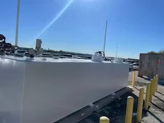 Low-angle view of a long fuel tank coated bright white, with yellow bollards and an open lot in the background.