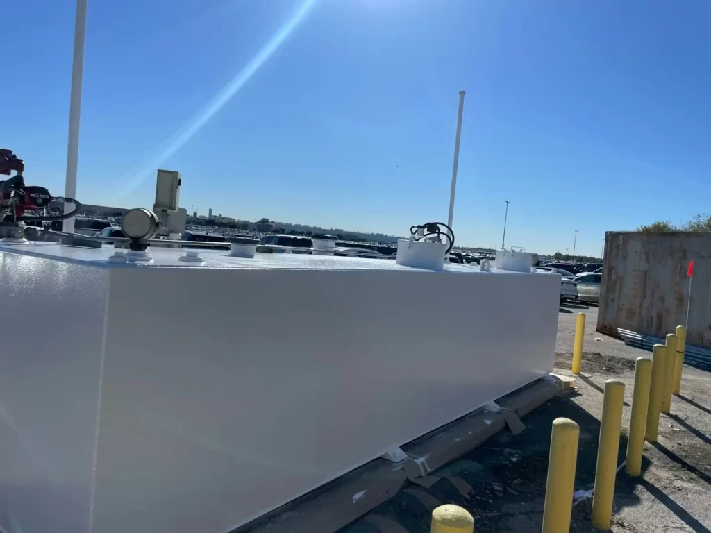 Glossy white-painted fuel tank seen from the side, with yellow bollards and a parking lot full of cars behind it.