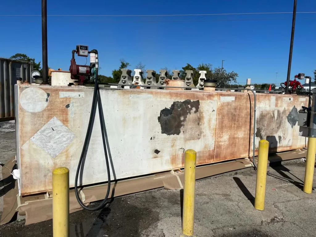 Side view of a heavily weathered fuel tank with a pump unit and hoses on top, surrounded by yellow bollards.