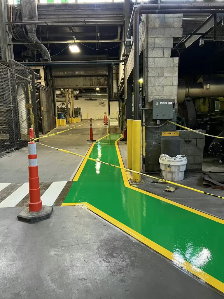 Industrial facility interior with a freshly painted green pedestrian path bordered in yellow, curving around columns and machinery; cones and caution tape block access.