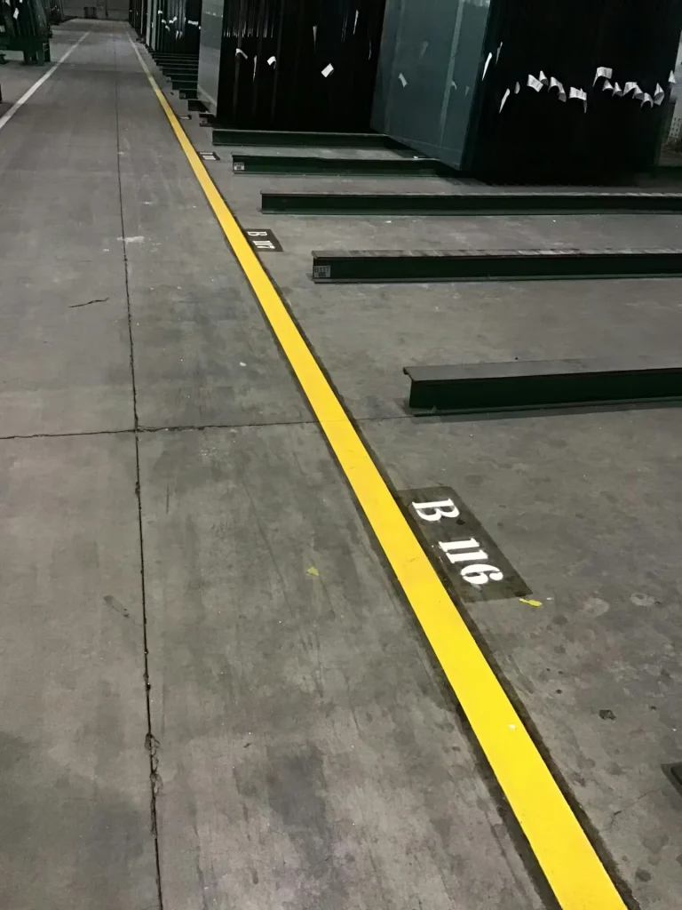 Warehouse aisle with a single bright yellow floor line marking a boundary beside stacked glass sheets on racks; stenciled bay labels (e.g., “B 116”) appear on the floor.
