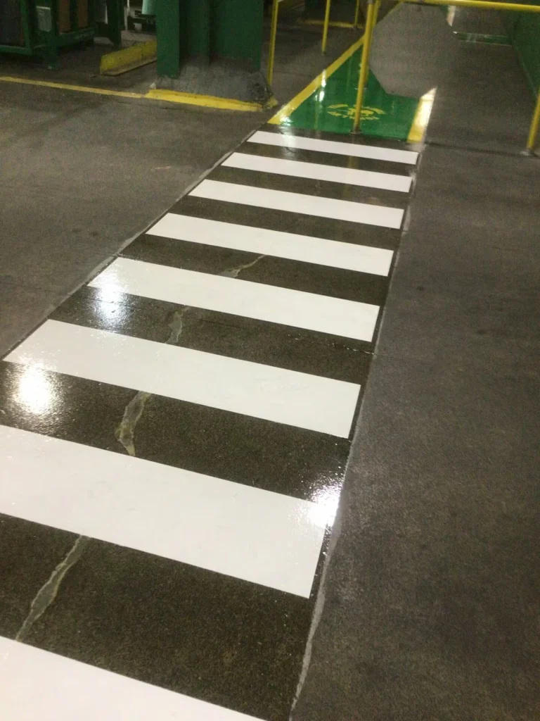 White zebra crosswalk leading toward stacked wrapped pallets and racking; orange cones and caution tape block the crossing, with a green lane visible at the bottom edge.