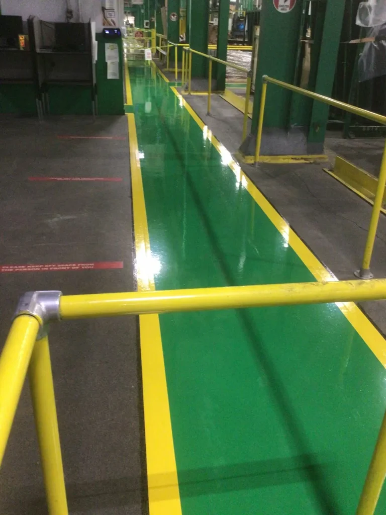 Narrow corridor in an industrial area with yellow guardrails flanking a shiny green walkway bordered in yellow; equipment and workstations line the sides.