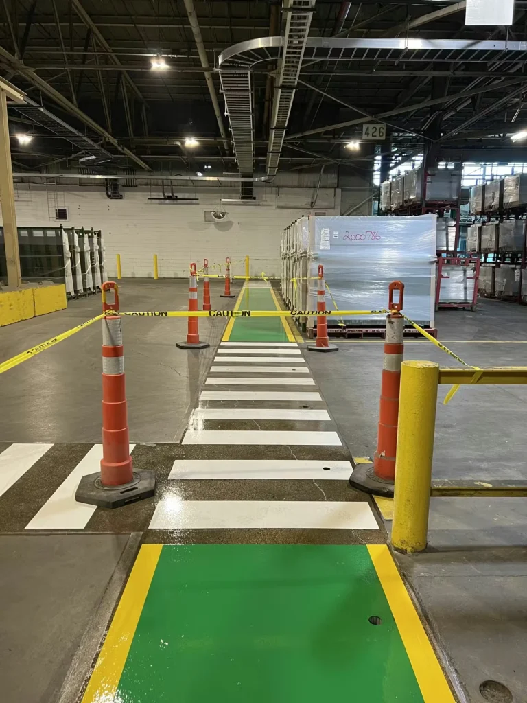 Long straight green walkway with yellow edges running beside a tall wire-mesh safety fence inside a warehouse; cones and caution tape mark the route ahead.